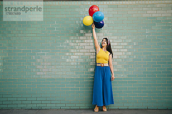 Frau mit erhobener Hand hält bunte Luftballons vor einer blaugrünen Ziegelwand