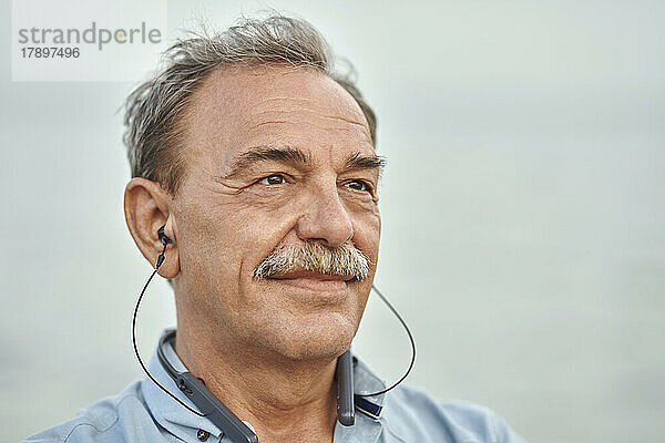 Nachdenklicher älterer Mann mit In-Ear-Kopfhörern  der vor dem Meer Musik hört