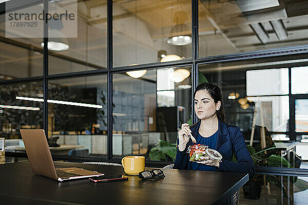 Junge Geschäftsfrau isst Obst und schaut im Büro auf den Laptop