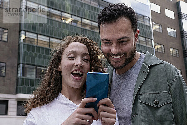 Glückliche Frau  die vor dem Gebäude ihr Smartphone mit einer Freundin teilt