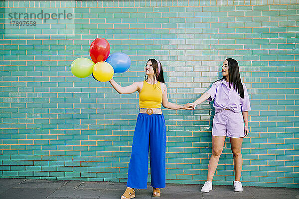 Frau hält Hand eines Freundes mit bunten Luftballons vor türkisfarbener Wand