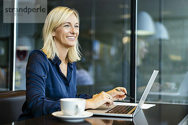 Lächelnde blonde Geschäftsfrau mit Laptop sitzt im Café