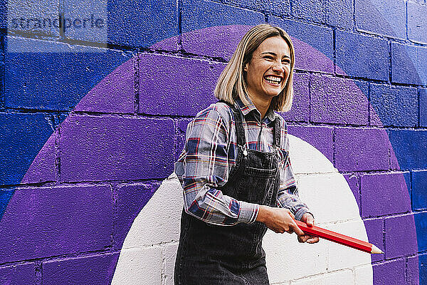 Fröhliche junge Frau hält großen Bleistift vor Regenbogenwandbild an der Wand