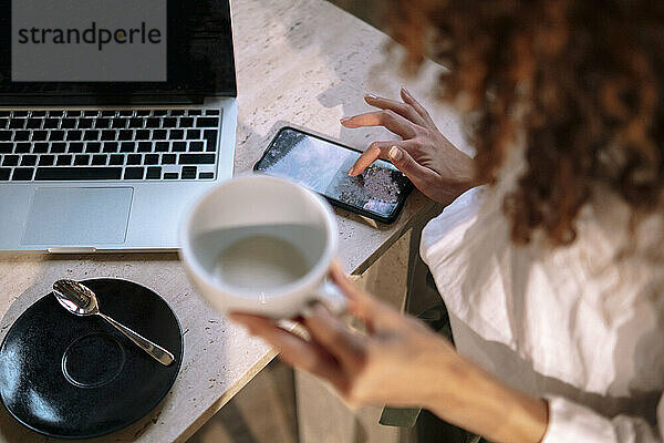 Hände einer Geschäftsfrau mit Kaffeetasse und Smartphone  die mit Laptop am Schreibtisch sitzt