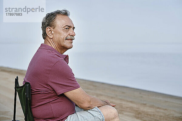 Nachdenklicher älterer Mann sitzt auf einem Klappstuhl am Strand