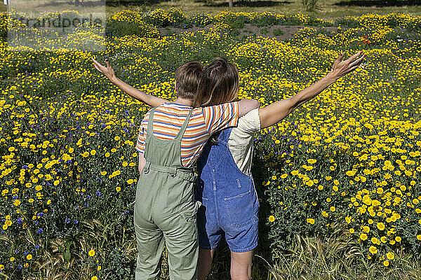 Nicht-binäre Person umarmt einen Freund  der mit ausgestreckten Armen vor blühenden Pflanzen steht