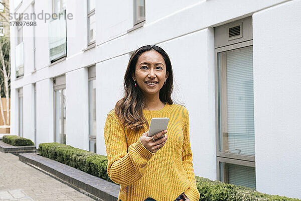 Happy young woman holding mobile phone by building