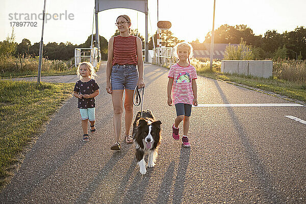 Mädchen und Mutter gehen bei Sonnenuntergang mit Hund auf der Straße spazieren