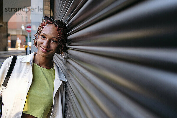 Lächelnde junge Frau mit Sonnenbrille lehnt an der Wand