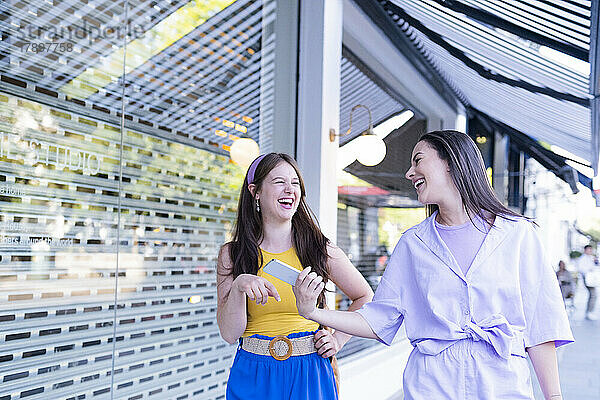 Fröhliche junge Frauen schauen sich an  während sie in der Nähe des Ladens spazieren gehen