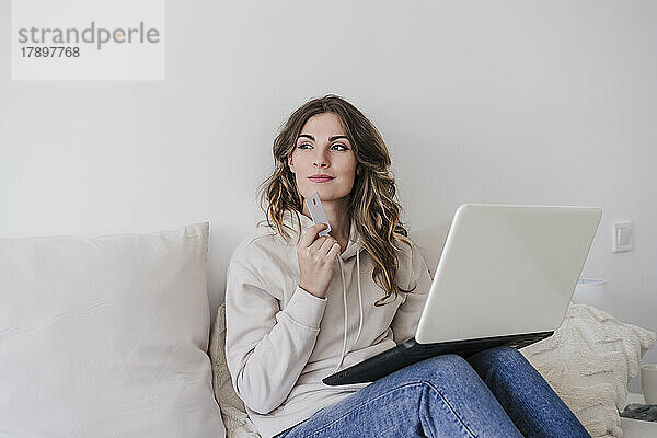 Nachdenkliche Frau mit Kreditkarte und Laptop sitzt zu Hause