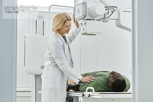Lächelnder Radiologe im Gespräch mit Patient  der in der Klinik auf dem Röntgengerät liegt