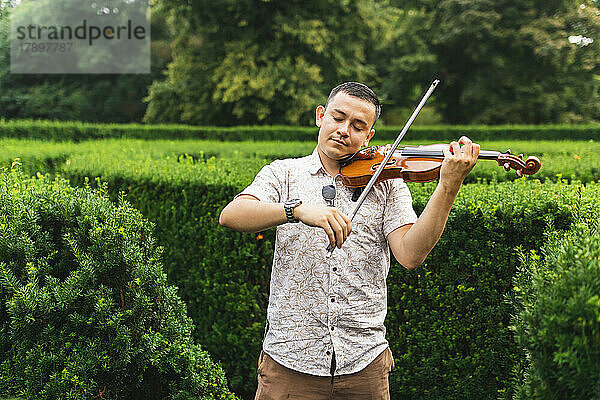 Junger Mann mit geschlossenen Augen spielt Geige neben Pflanzen im Park