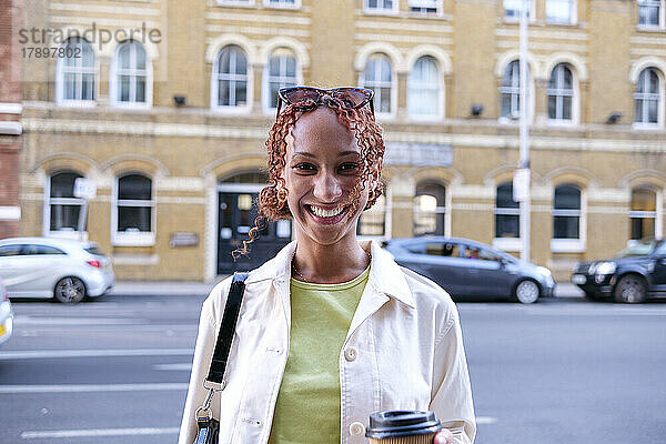 Glückliche junge Frau mit lockigem Haar hält eine Einweg-Kaffeetasse in der Hand