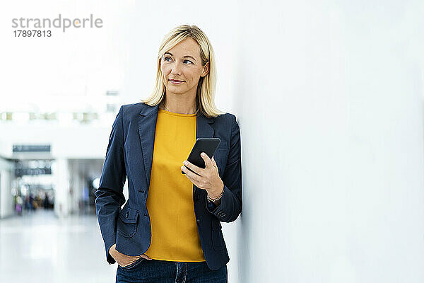 Nachdenkliche Geschäftsfrau mit Mobiltelefon an der Wand