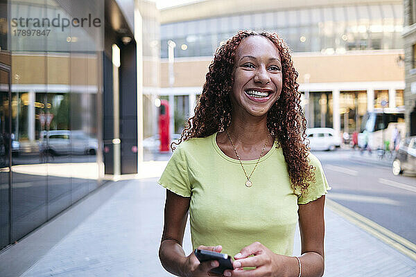 Glückliche junge Frau hält ihr Mobiltelefon auf dem Fußweg
