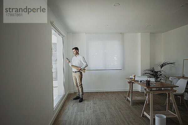 Architekt hält aufgerollten Bauplan mit Smartphone in der Hand und steht am Fenster im Büro