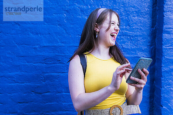 Fröhliche Frau hält Mobiltelefon vor blauer Wand
