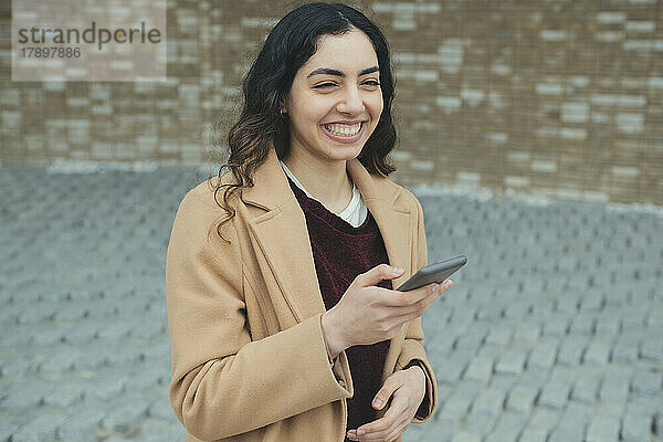 Glückliche junge Frau mit Smartphone  die an der Wand steht