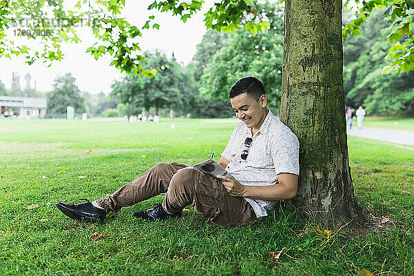 Lächelnder Mann liest ein Buch und lehnt an einem Baumstamm im Park
