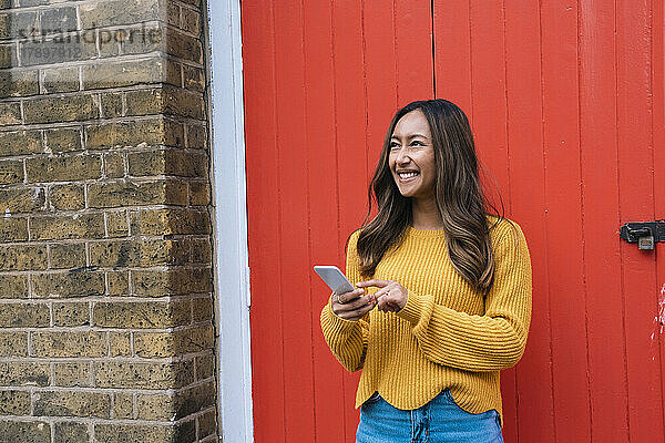 Happy young woman with mobile phone standing in front of red door