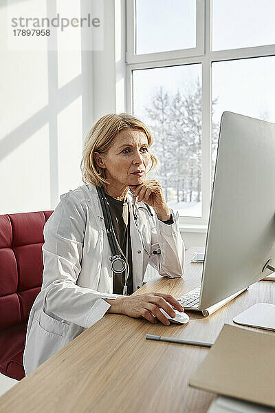 Konzentrierter Arzt arbeitet am Desktop-PC und sitzt am Schreibtisch in der Klinik