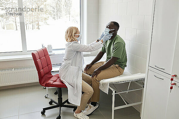 Arzt untersucht Patienten mit schützender Gesichtsmaske  der in der Klinik auf dem Bett sitzt