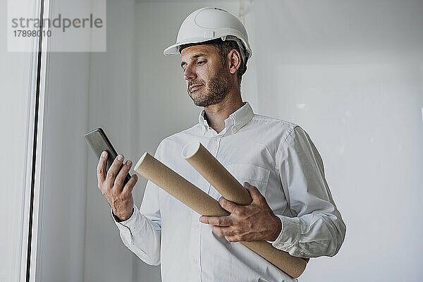 Reifer Ingenieur hält zusammengerolltes Papier in der Hand und blickt auf sein Smartphone vor einer weißen Wand