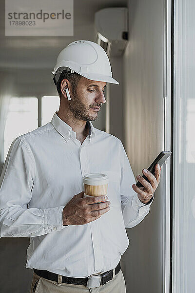 Ingenieur hält Einwegbecher in der Hand und schaut im Büro auf sein Mobiltelefon