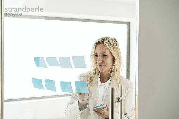 Businesswoman sticking adhesive notes on glass wall in office