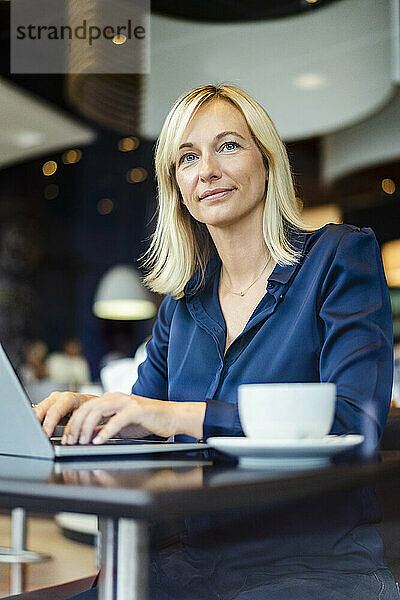 Lächelnde Geschäftsfrau mit Laptop sitzt am Tisch im Café