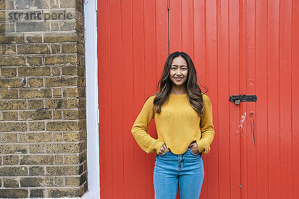 Smiling young woman with hands in pockets standing in front of red door