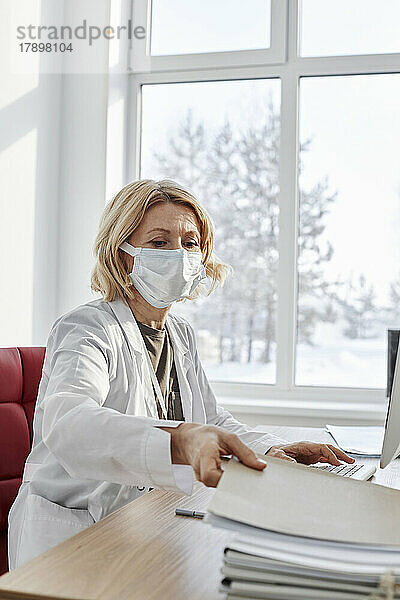 Arzt mit schützender Gesichtsmaske holt Akte am Schreibtisch in der Klinik ab