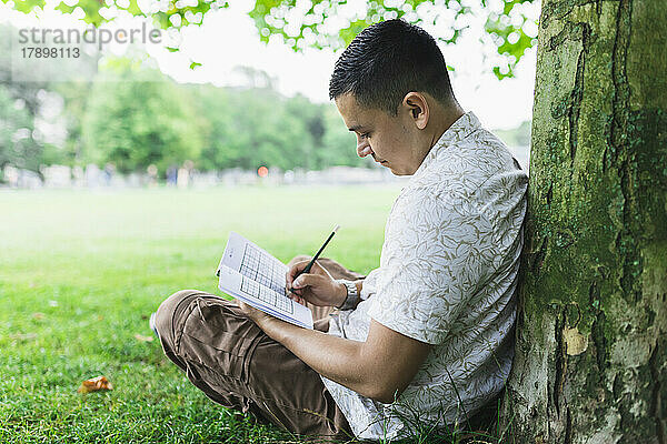 Mann  der am Baumstamm im Park sitzt und Sudoku in einem Buch löst