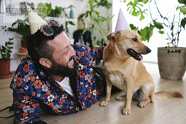 Glücklicher Mann mit Partyhut und Sonnenbrille  der zu Hause neben dem Hund liegt