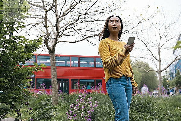 Nachdenkliche junge Frau mit Mobiltelefon steht im Park