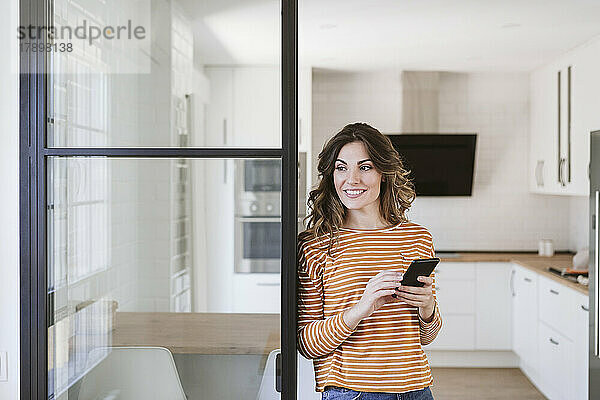 Smiling young woman holding mobile phone at a glass wall in kitchen