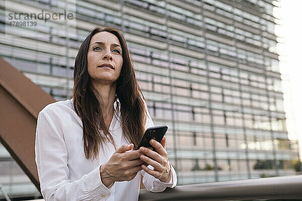 Nachdenkliche Geschäftsfrau mit Telefon steht am Geländer auf der Brücke