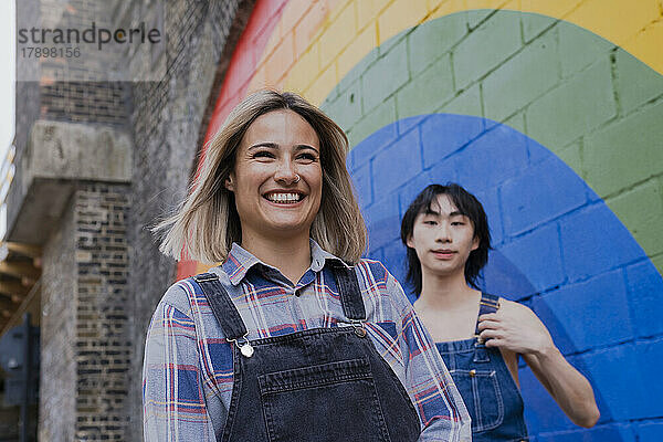 Glückliche junge Frau  die mit einem Freund am an der Wand gemalten Regenbogen spazieren geht