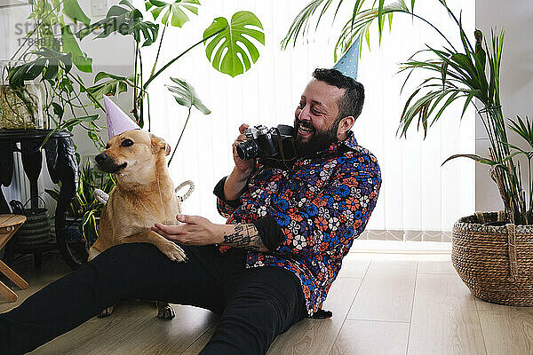 Fröhlicher Mann fotografiert Hund mit Partyhut zu Hause