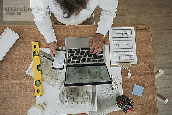 Architect holding smart phone sitting with laptop at desk in office