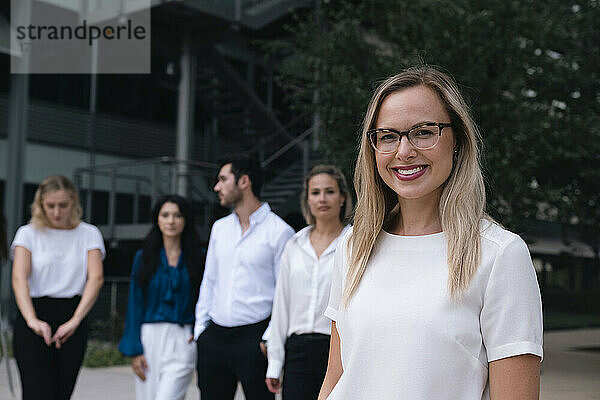 Glückliche Geschäftsfrau mit Brille steht vor Kollegen im Büropark