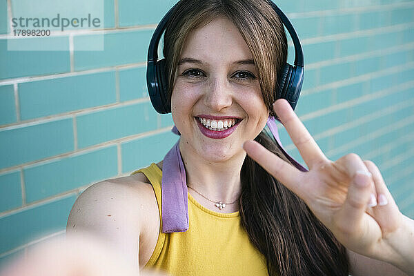 Glückliche Frau mit Kopfhörern zeigt ein Friedenszeichen an einer blaugrünen Wand
