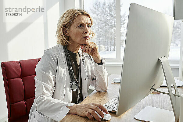 Arzt mit Hand am Kinn und Desktop-PC am Schreibtisch in der Klinik