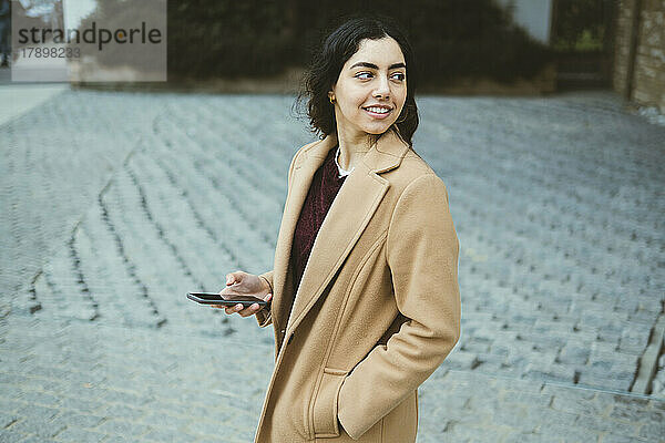 Lächelnde junge Frau im Trenchcoat mit Smartphone