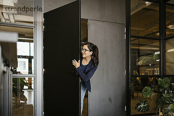 Lächelnde junge Geschäftsfrau späht durch die Tür im Büro
