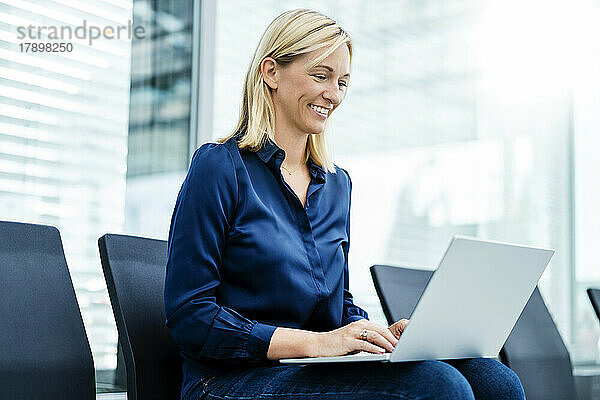 Lächelnde Geschäftsfrau sitzt mit Laptop auf einem Stuhl in der Lobby