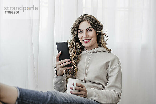 Lächelnde schöne Frau mit Kaffeetasse und Mobiltelefon sitzt zu Hause