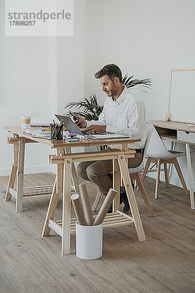 Reifer Architekt hält Smartphone und Laptop am Schreibtisch