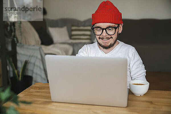 Lächelnder Mann mit Strickmütze und Laptop zu Hause